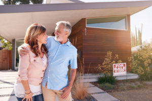 a senior couple stands in front of their home that is for sale